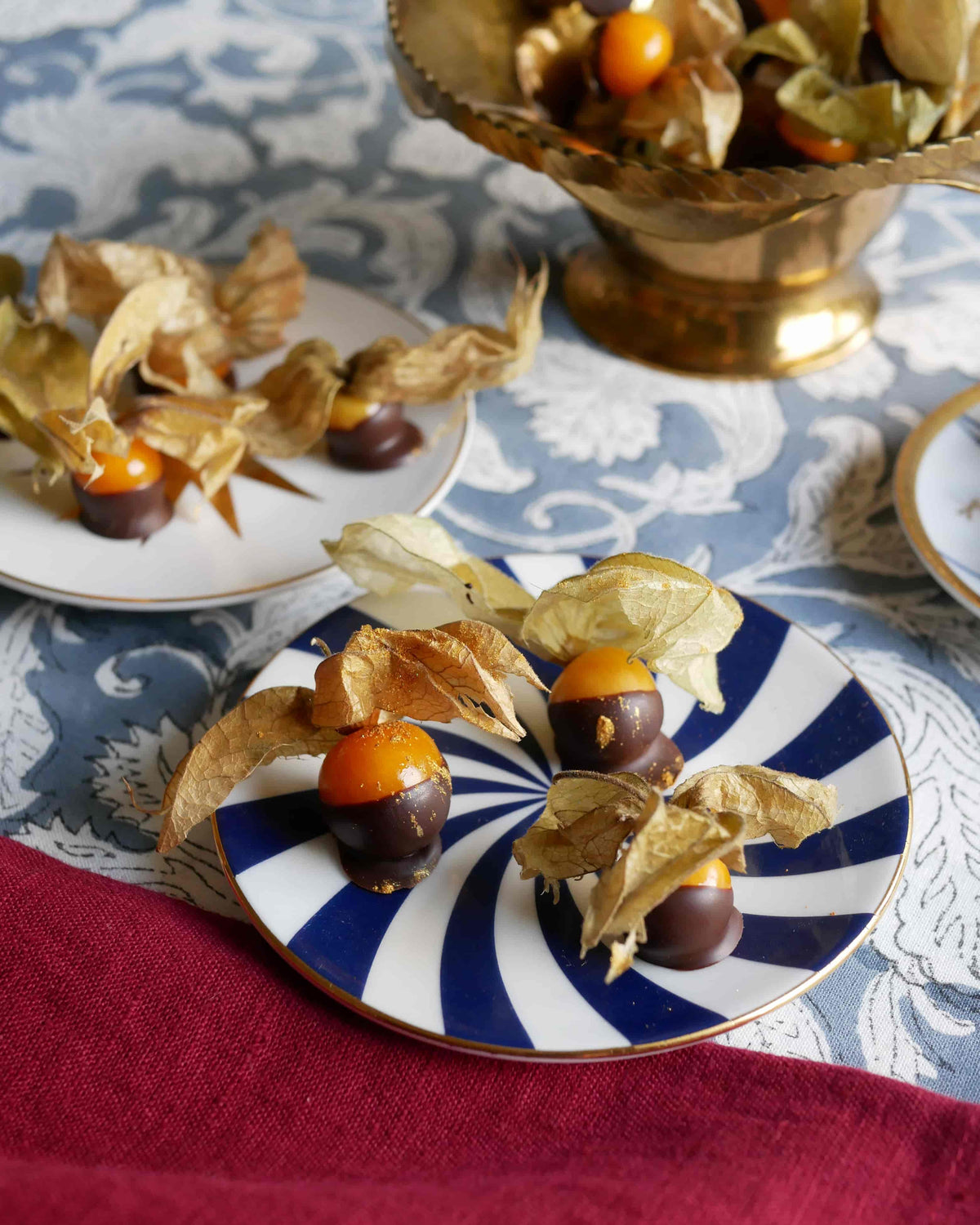 Schokolierte Physalis mit essbarem Gold auf blau-weißem Porzellanteller, arrangiert auf der Weihnachtstafel „Blue Velvet“ mit graublauer Blockprint-Tischdecke und weinrotem Leinen.