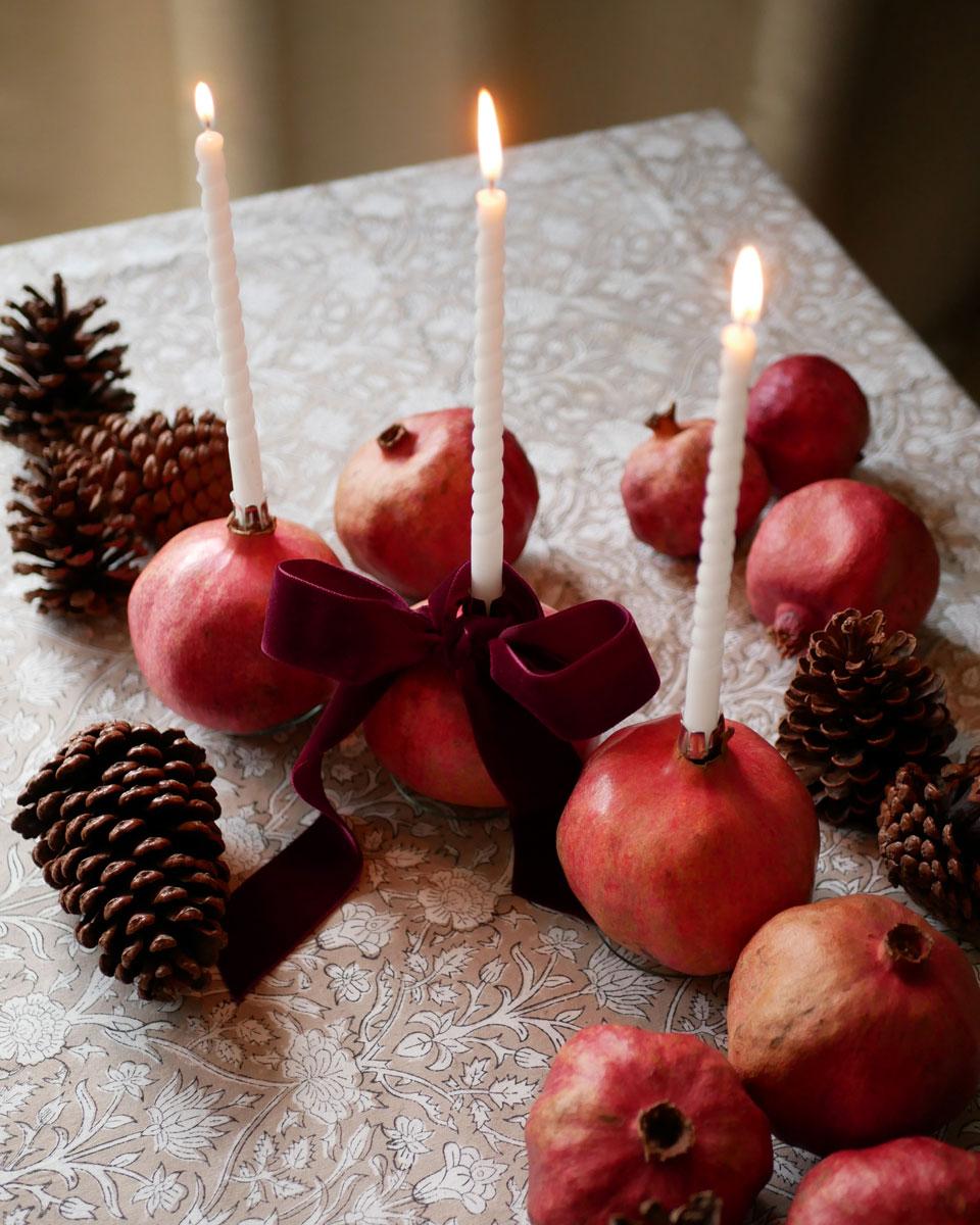 Granatapfel-Kerzenhalter mit weißen Baumkerzen und bordeauxfarbener Samtschleife auf einer festlichen Weihnachtstafel mit sandfarbener Blockprint-Tischdecke und Tannenzapfen.